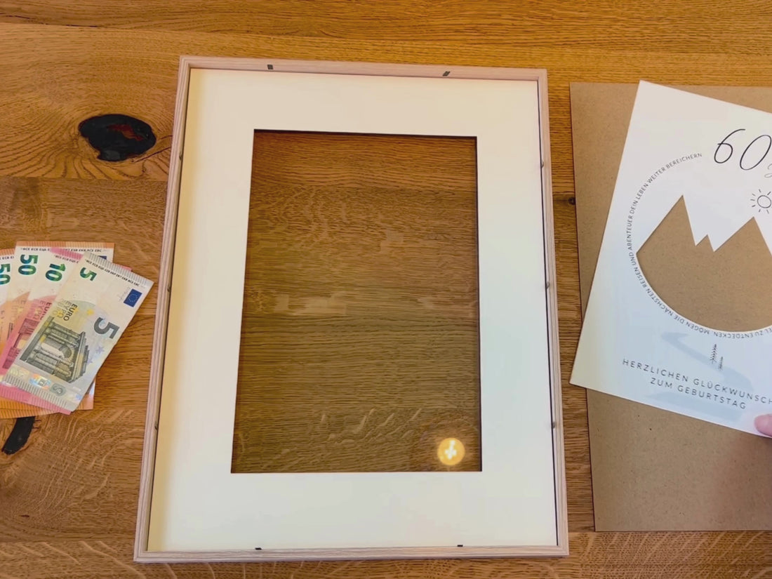 Videoanleitung: So funktioniert die Geldgeschenk-Vorlage mit Bergmotiv. Gezeigt wird, wie die ausgeschnittene Vorlage in einen Bilderrahmen gelegt, Geldscheine dahinter platziert und das fertige Geschenk dekorativ präsentiert wird.